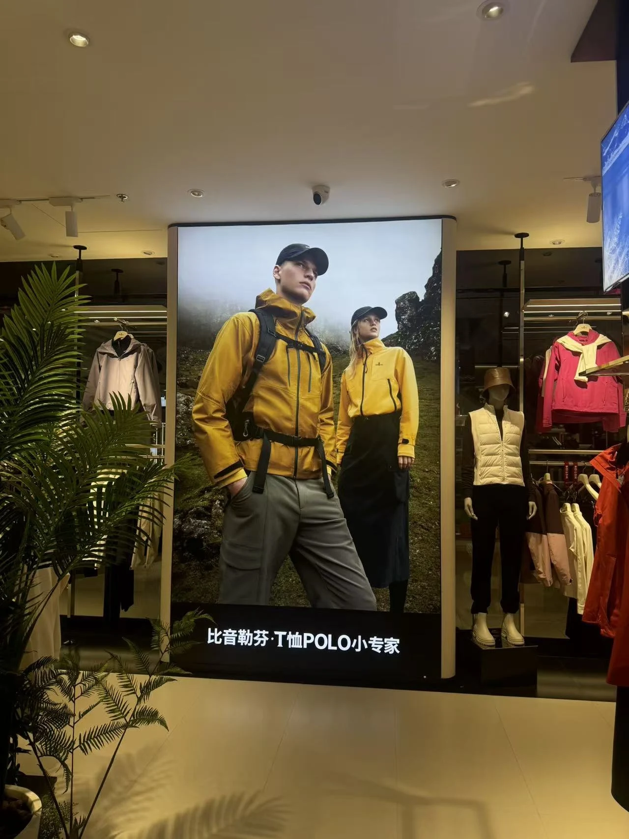 Portrait fabric lightbox between clothing racks inside the Biemlofen store, showing two models in outdoor jackets
